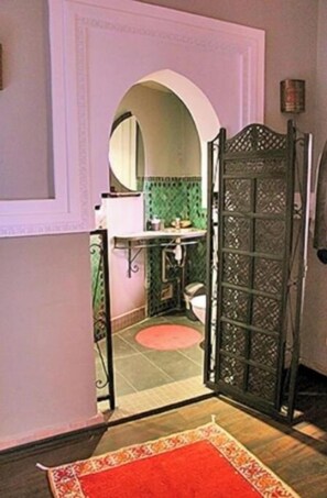 Una puerta abierta que da a un baño con pared de azulejos verdes, un lavabo blanco y un espejo.