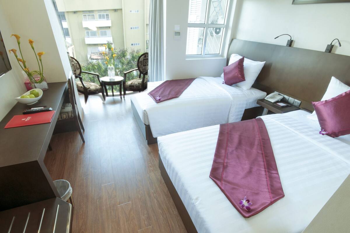 Ein Hotelzimmer mit zwei Betten, einem Schreibtisch mit Blumenvase, einem Sessel und Blick ins Freie.