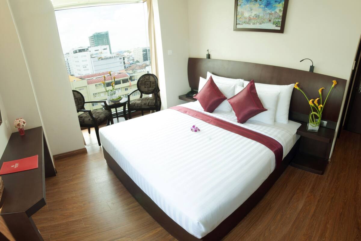 Ein Hotelzimmer mit einem großen Bett, einem Schreibtisch, einem Sessel, einem kleinen Tisch und einem Fenster mit Blick auf die Stadt.
