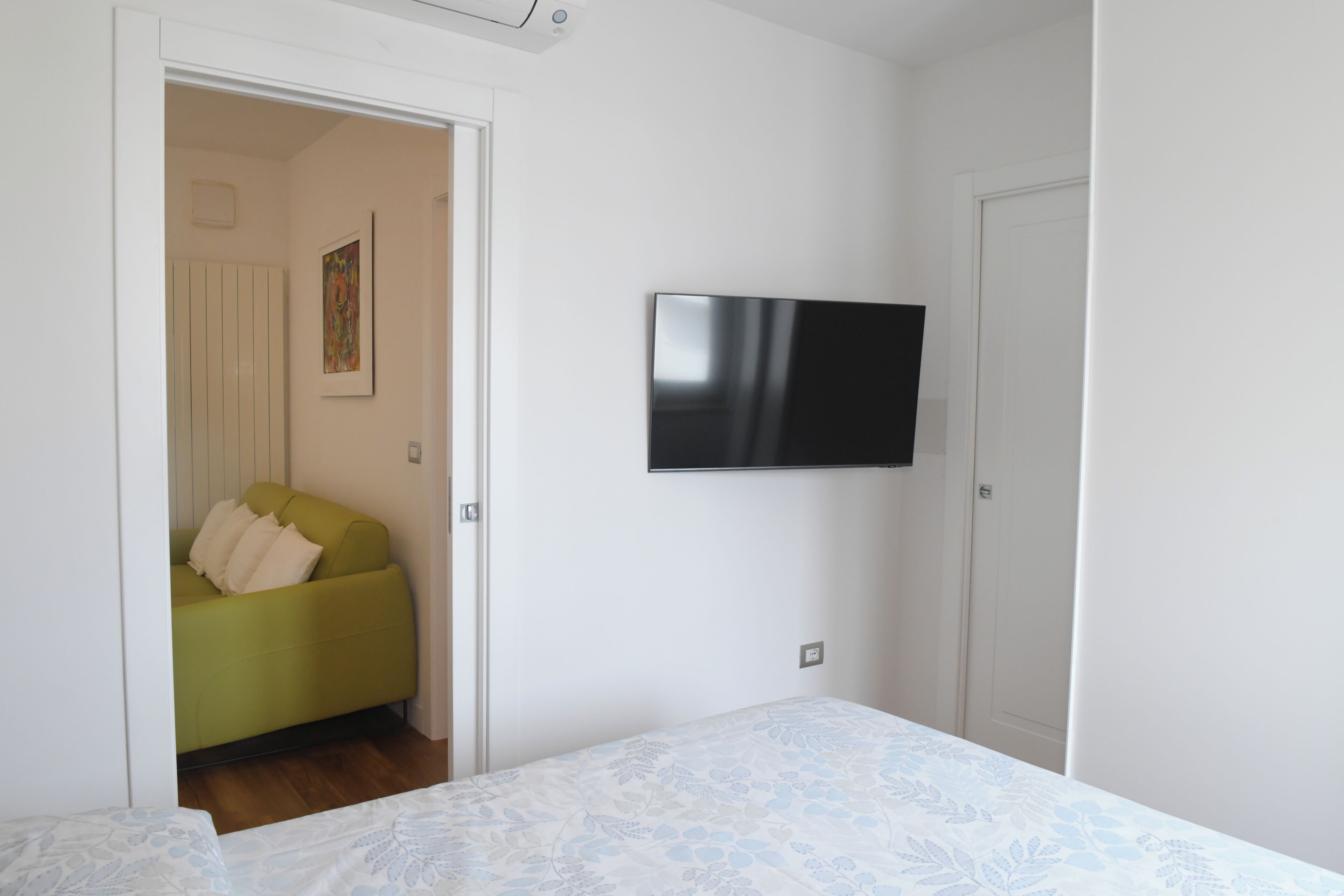 Ein Schlafzimmer mit einem Bett, einem grünen Sofa, einem an der Wand montierten Flachbildfernseher und einer Tür, die in einen anderen Raum führt.