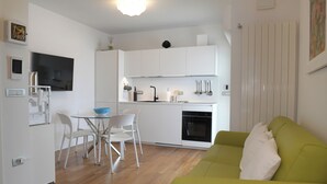 A modern kitchen with white cabinets, a dining table with chairs, a green sofa, and a flat-screen TV mounted on the wall. - Lecasinedigiò2 Cozy and refined apartment with sea view terrace (Alghero)