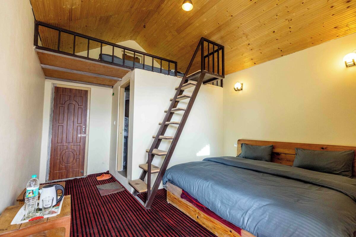 Une chambre avec un plafond en bois, un lit avec une literie grise, une échelle en bois menant à une mezzanine et une porte.
