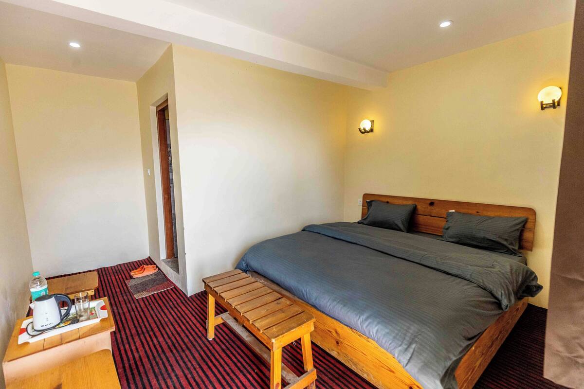 Une chambre à coucher avec un lit, un banc en bois, une petite table avec une bouilloire, des verres et une bouteille d’eau.