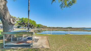 A picnic table with a metal frame and a bench, a large tree, and a body of water with a dock in the background.