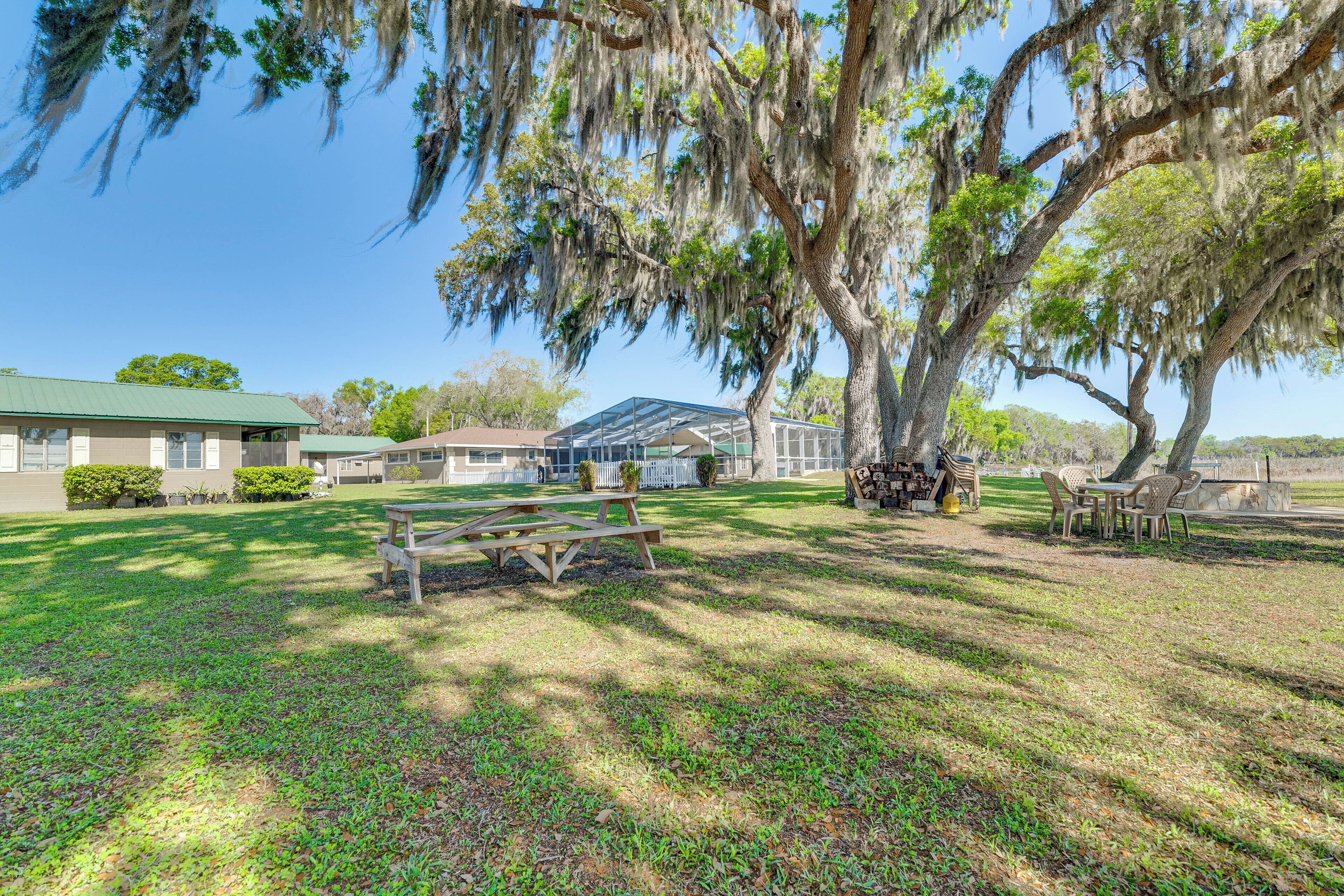 A grassy area with a picnic table and chairs, a large tree with hanging moss, and a building with a green roof in the background.