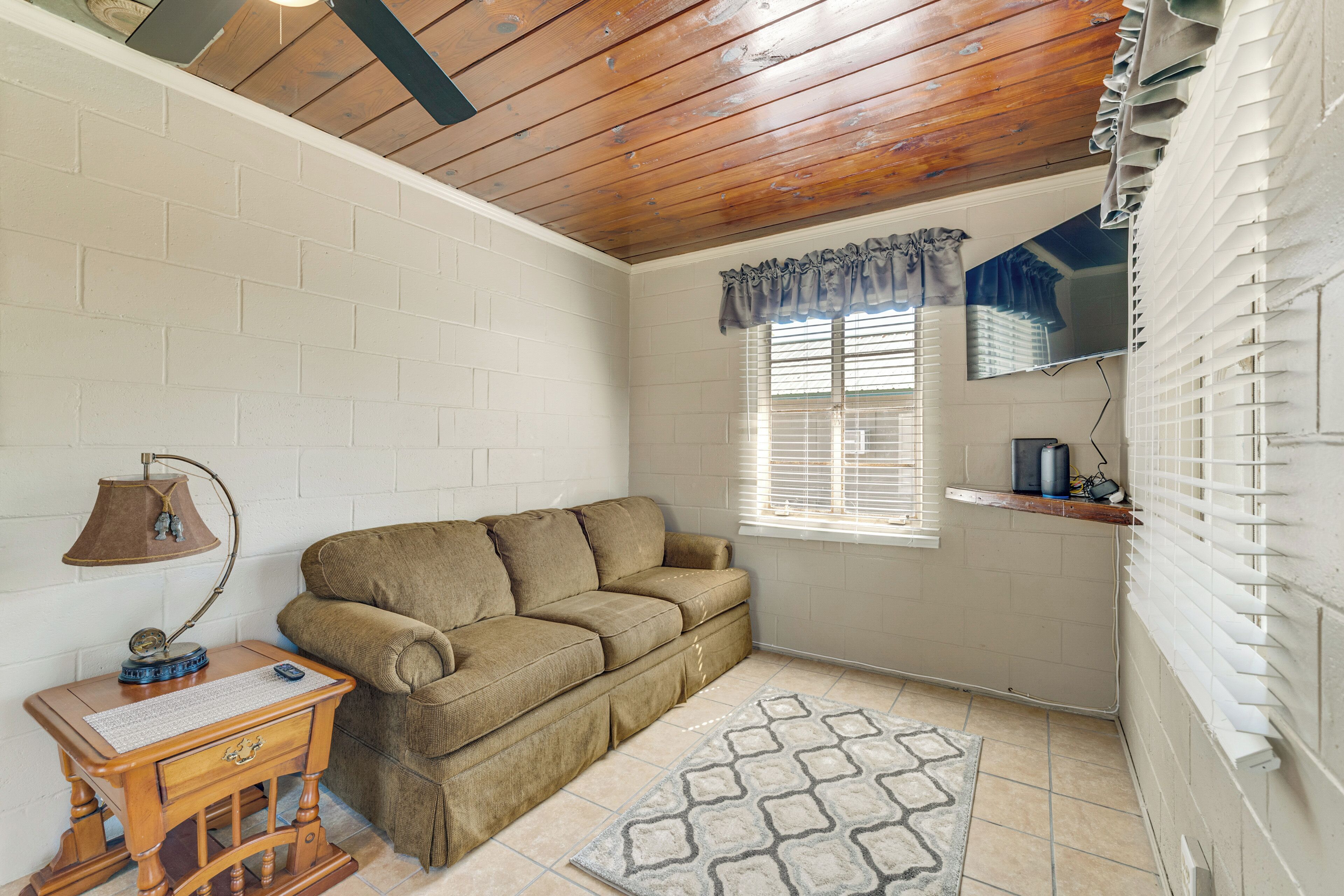 A room with a wooden ceiling, a brown sofa, a small wooden table with a lamp, a window with blinds, and a patterned rug.