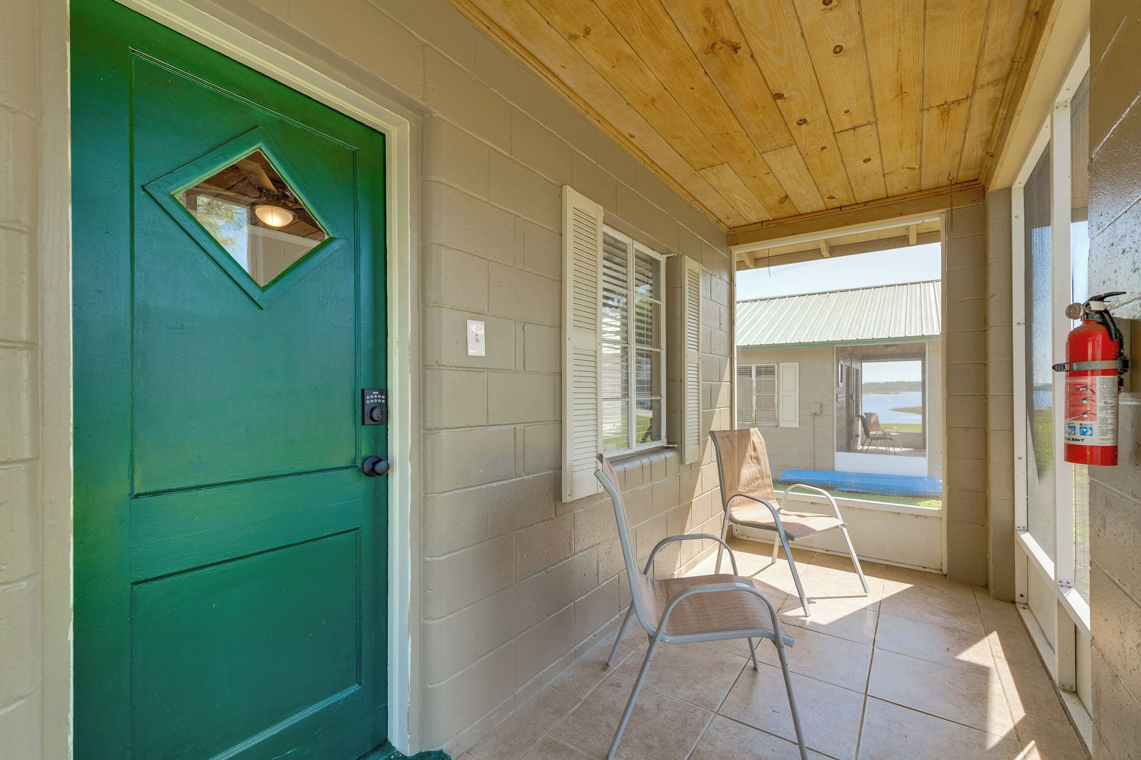 A green door with a small window, white shutters, a fire extinguisher, and a chair in a covered porch area.