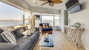 A living room with a sofa, a coffee table, a ceiling fan, a TV mounted on the wall, and a view of the water outside. - Waterfront Resort Home! Extended Jensen Beach Stay (Jensen Beach)