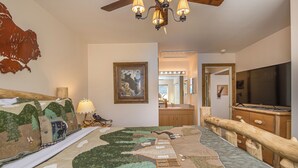 A bedroom with a large bed, a wooden headboard, a ceiling fan, a mounted TV, and a bathroom visible through an open door.
