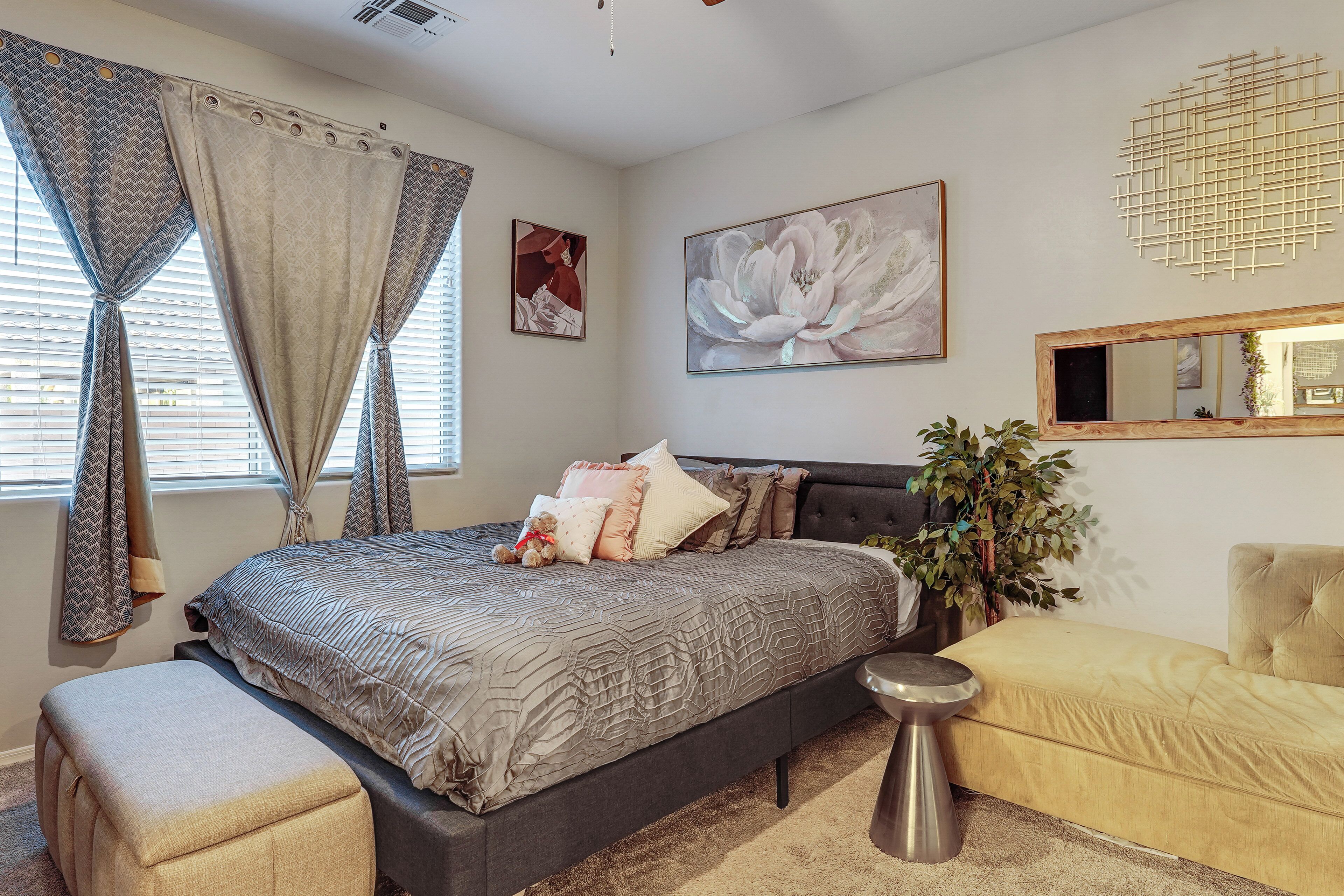 A bedroom with a bed, a bedside table, a chair, a large window with curtains, and a wall-mounted mirror.