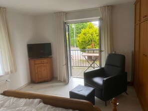 Ein Schlafzimmer mit einem Bett, einem Fernseher auf einer Holzkommode, einem schwarzen Sessel, einem kleinen Tisch und einer Glastür, die zu einem Außenbereich mit Tisch und Stühlen führt.