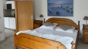 Une chambre à coucher avec un lit en bois, un tableau représentant une scène de plage, un coin cuisine et une table de chevet en bois.