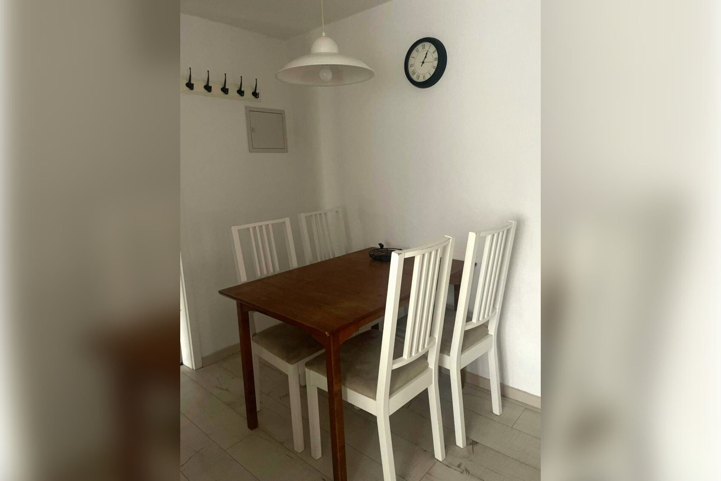 Un comedor con una mesa de madera y sillas blancas, una lámpara colgante blanca y un reloj de pared.