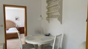 Ein Zimmer mit rundem Tisch, zwei Stühlen, einem Bett mit Kopfteil, einem Nachttisch mit Lampe und einem an der Wand befestigten Regal mit Gläsern.