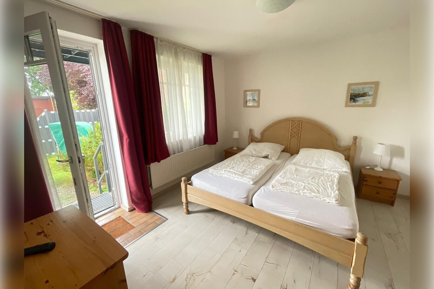 A double bed with a wooden headboard, a bedside table with a lamp, a window with curtains, and a view of the outside.