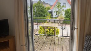 A balcony with a wooden table and chairs, a metal railing, and a view of a white house with a red roof.