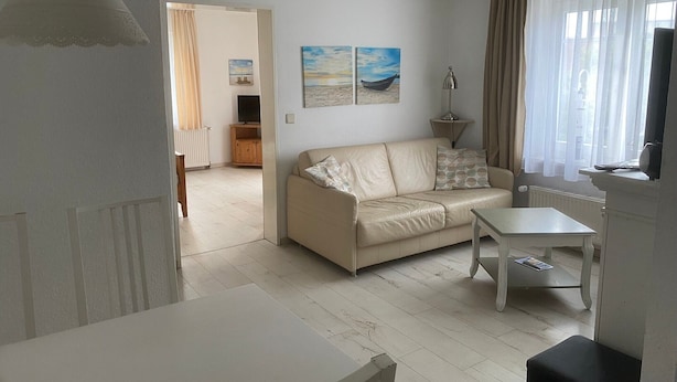 A living room with a beige sofa, a small table, and a TV. There are two paintings on the wall, a lamp, and a window with curtains.