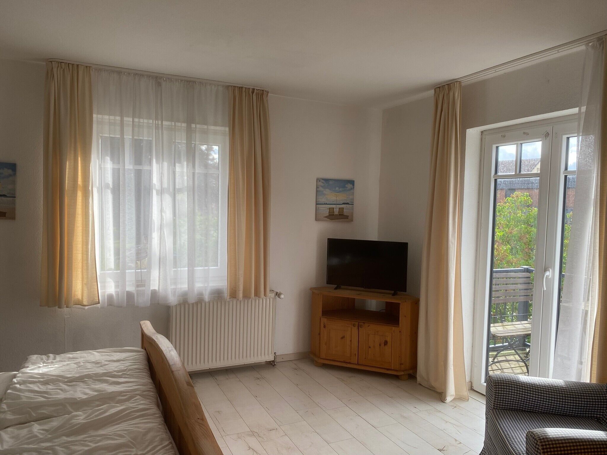 A bedroom with a bed, a TV on a wooden stand, a radiator, and a balcony with a view of greenery.