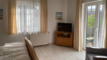 A bedroom with a bed, a TV on a wooden stand, a radiator, and a balcony with a view of greenery.