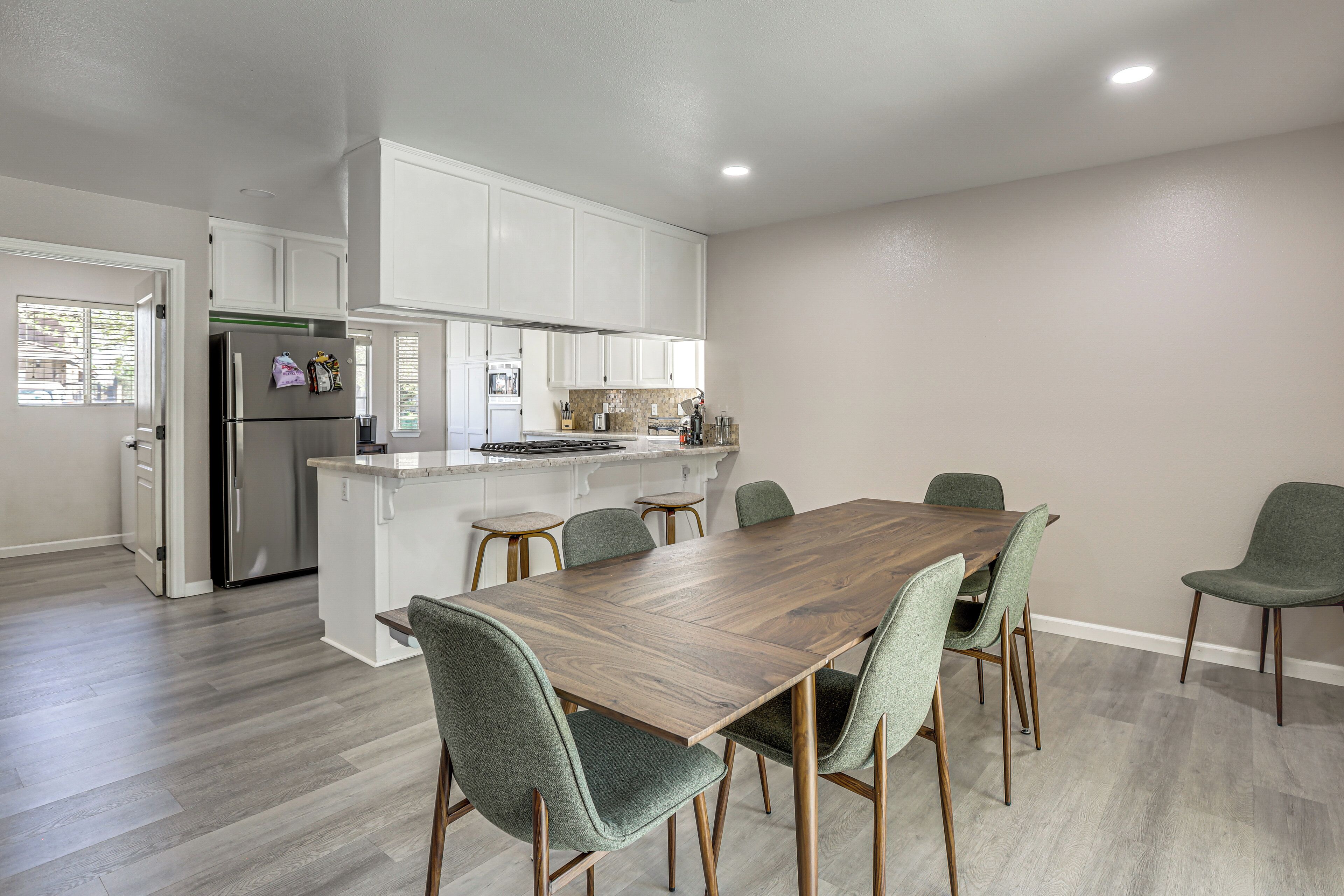 A modern kitchen with white cabinets, a stainless steel refrigerator, a wooden dining table with six chairs, and a small island with a sink.