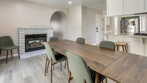 A dining area with a long wooden table, green upholstered chairs, a brick fireplace, and a kitchen with white cabinets.
