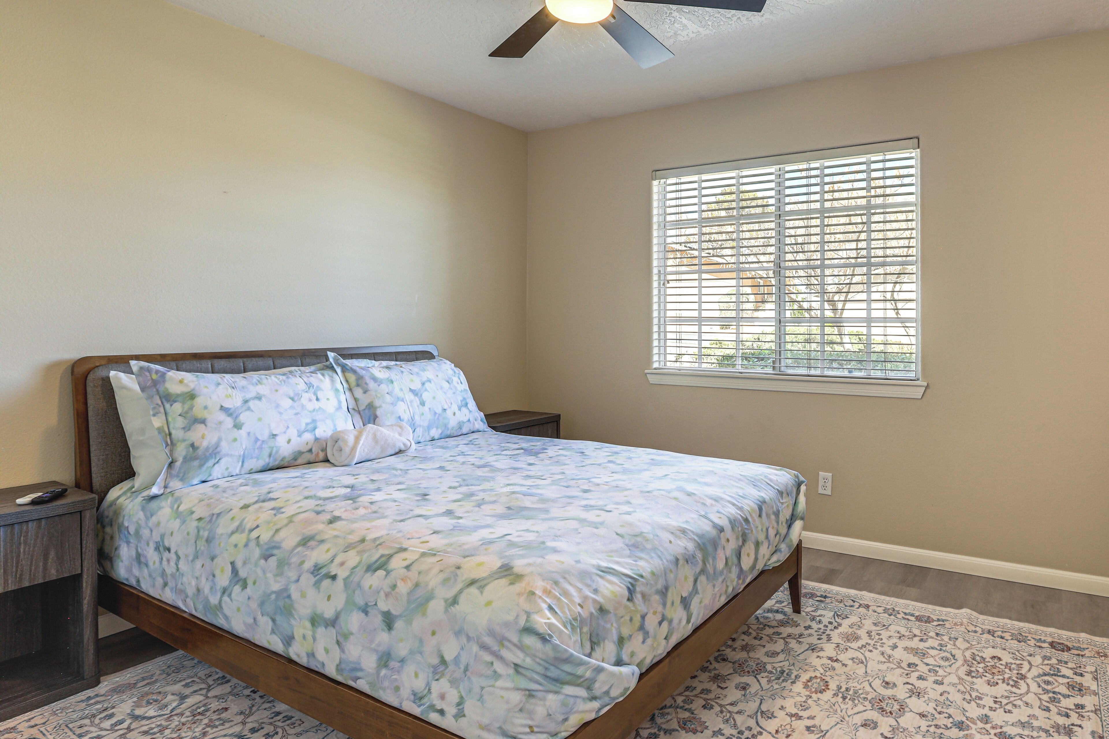 Une chambre à coucher comprenant un lit, une table de chevet, un ventilateur de plafond, une fenêtre avec des stores et un tapis à motifs.