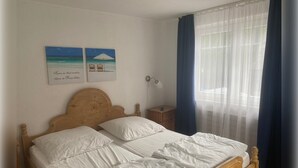 Un lit double avec du linge de lit blanc, une tête de lit en bois, une table de chevet avec une lampe et deux œuvres d’art murales représentant une scène de plage.