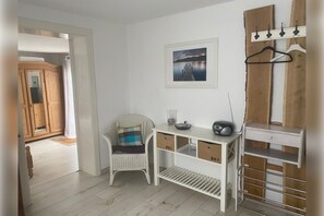 Ett rum med ett vitt skrivbord, en flätad stol, en tavla på väggen och en klädhängare.