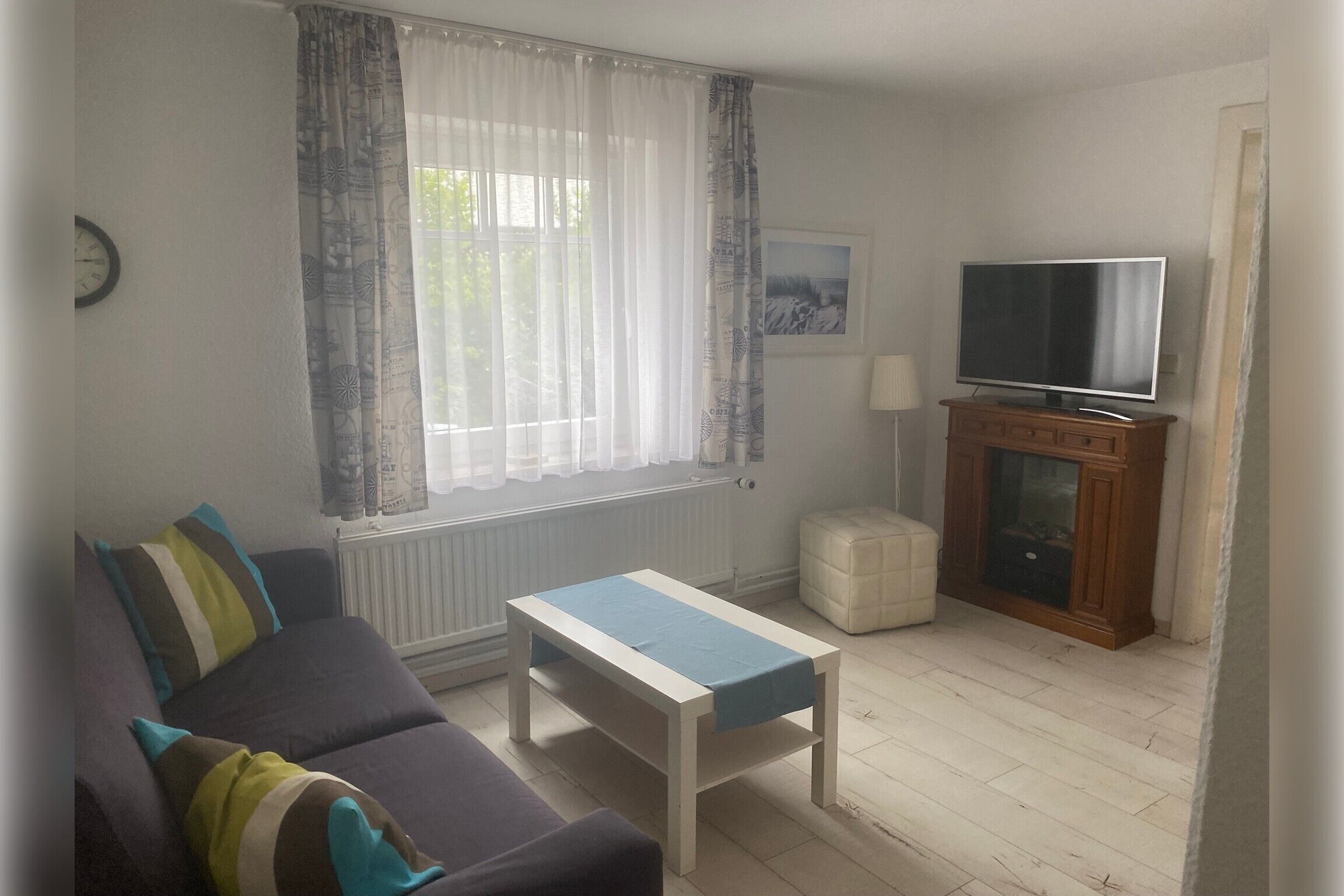 A living room with a sofa, a coffee table, a television on a cabinet, a window with curtains, and a radiator.