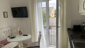 Una habitación con un sofá blanco, una mesa de vidrio, un televisor montado en la pared, un ventanal corredero al exterior y una zona de kitchenette.