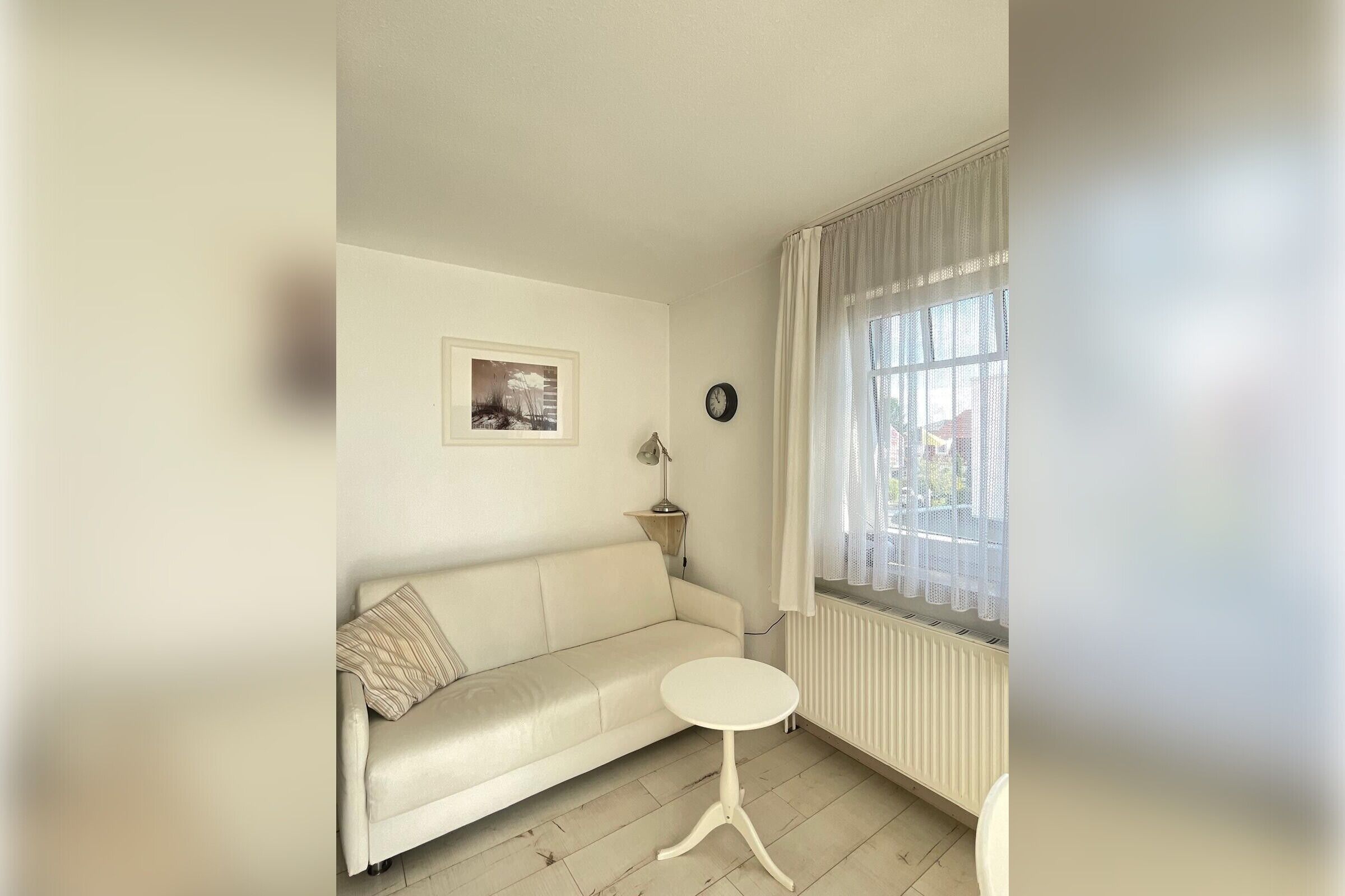 Ein Wohnzimmer mit einem weißen Sofa, einem kleinen runden Tisch, einer Lampe, einer Uhr und einem Fenster mit Vorhängen.