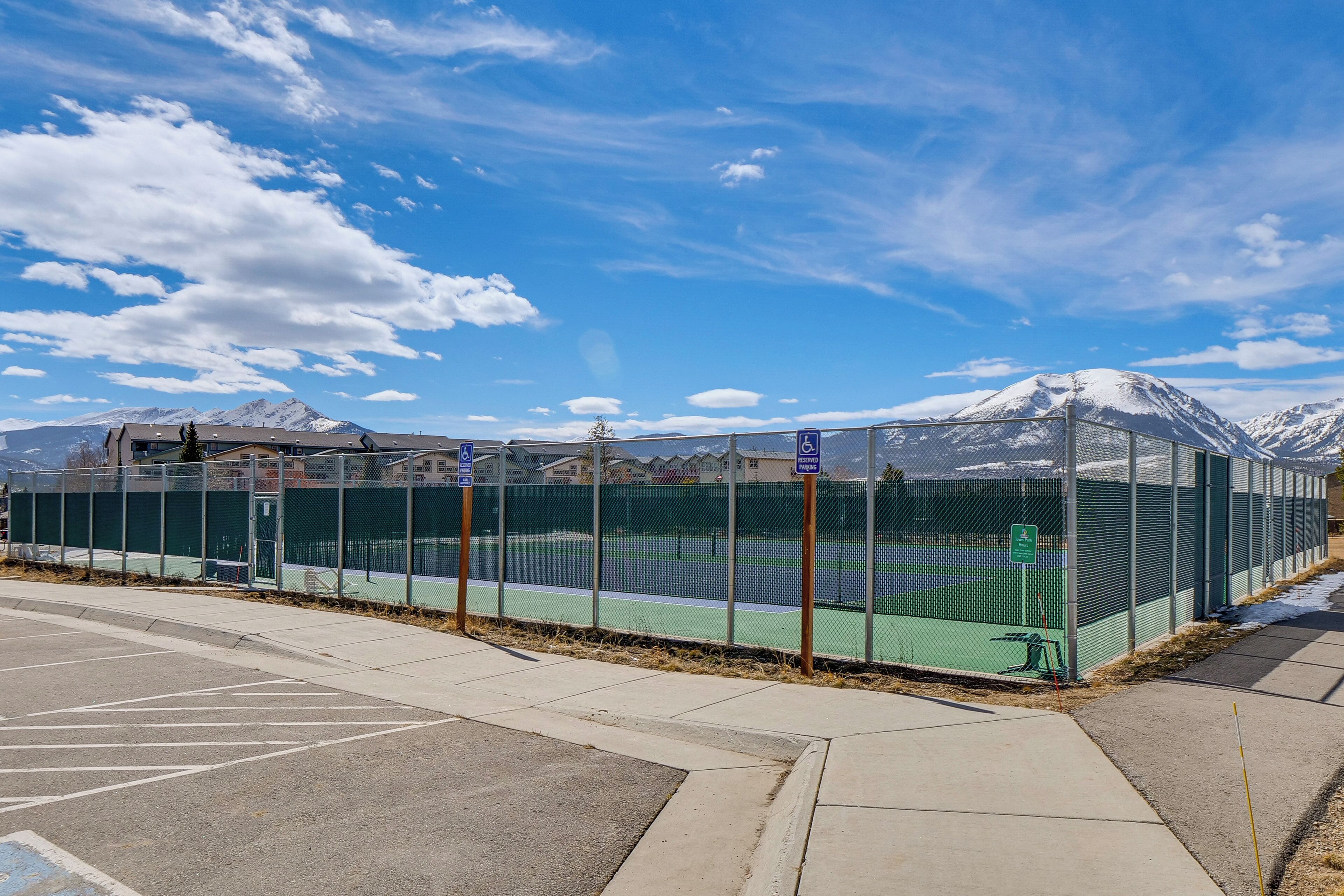 A tennis court with a fence, a parking lot, and a mountain in the background.