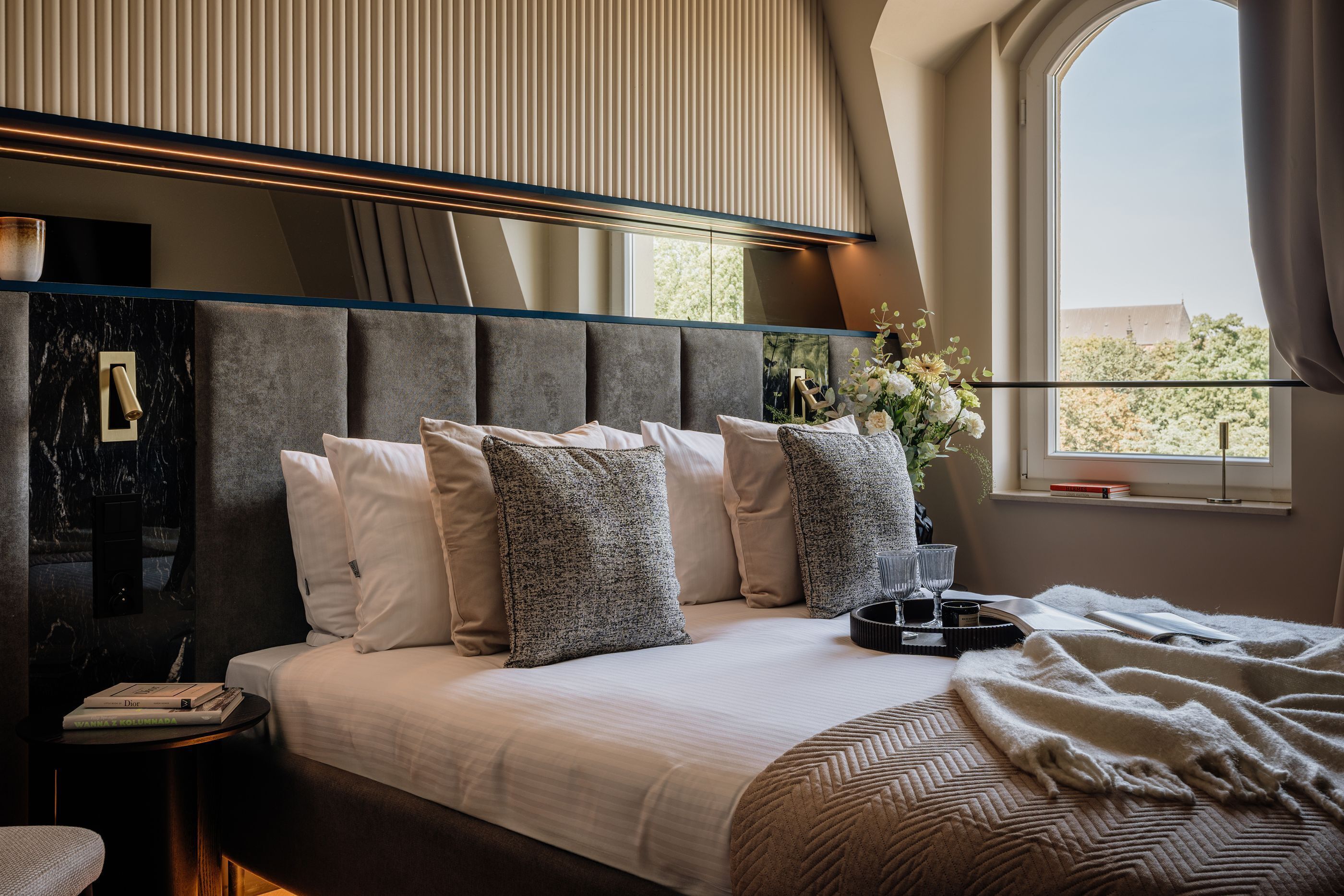 A modern hotel room with a large bed, a side table, a window with curtains, and a view of the outside.