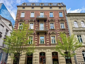 A multi-story building with a classic architectural style, featuring a central balcony with ornate railings, large windows, and a ground-level entrance with a sign.