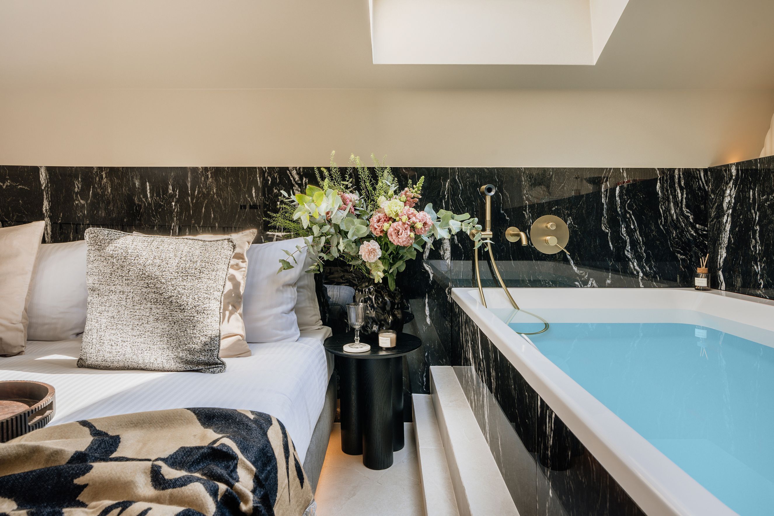 A bedroom with a bed, a bedside table with a vase of flowers, and a bathtub with a marble wall.