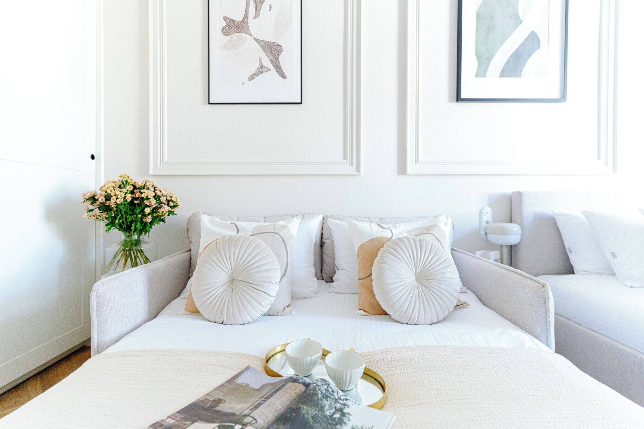A neatly arranged bedroom with a bed, two sofas, a vase with flowers, and framed artwork on the wall.