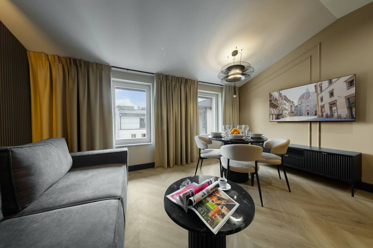 Una habitación de hotel moderna con sofá, mesa de comedor, sillas, una revista sobre una mesita y un ventanal con cortinas.