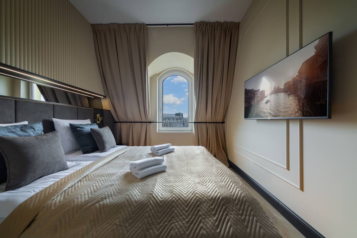 Habitación de hotel con una cama grande, televisor en la pared, ventana con cortinas y vista a la ciudad.