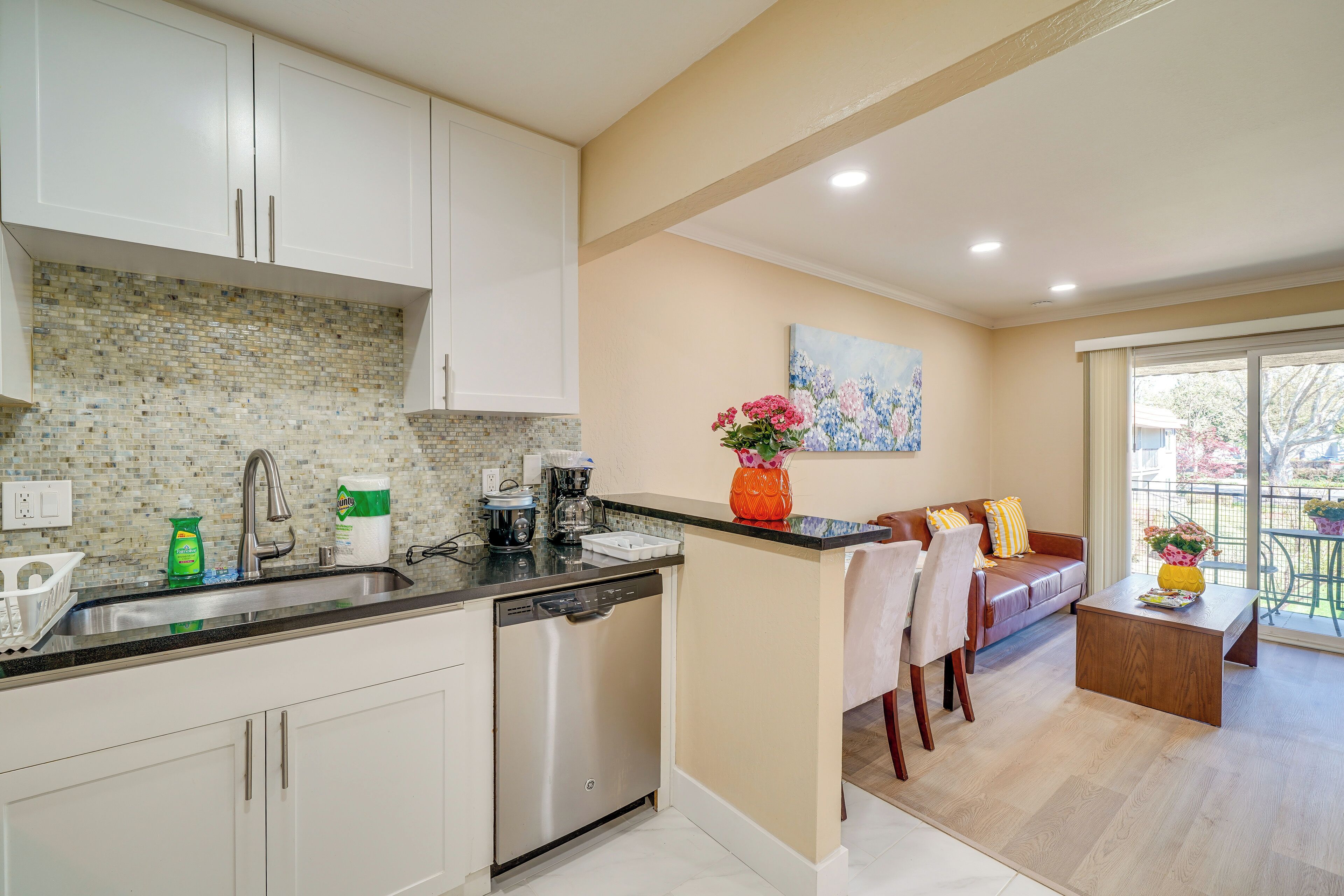 A modern kitchen with white cabinets, a dishwasher, and a coffee maker. There is a dining area with a sofa and a coffee table.