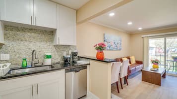 A modern kitchen with white cabinets, a dishwasher, and a coffee maker. There is a dining area with a sofa and a coffee table.
