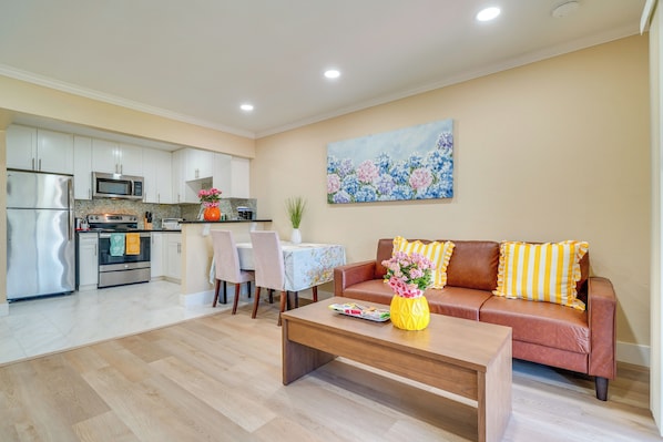 A modern kitchen with stainless steel appliances, a dining area with a table and chairs, a living room with a sofa and coffee table, and a floral painting on the wall.