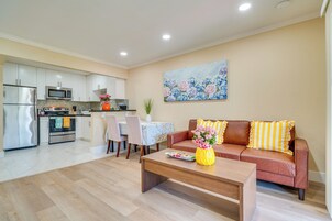 Una cocina moderna con electrodomésticos de acero inoxidable, un comedor con mesa y sillas, una sala de estar con sofá y mesa de centro, y un cuadro de flores en la pared.