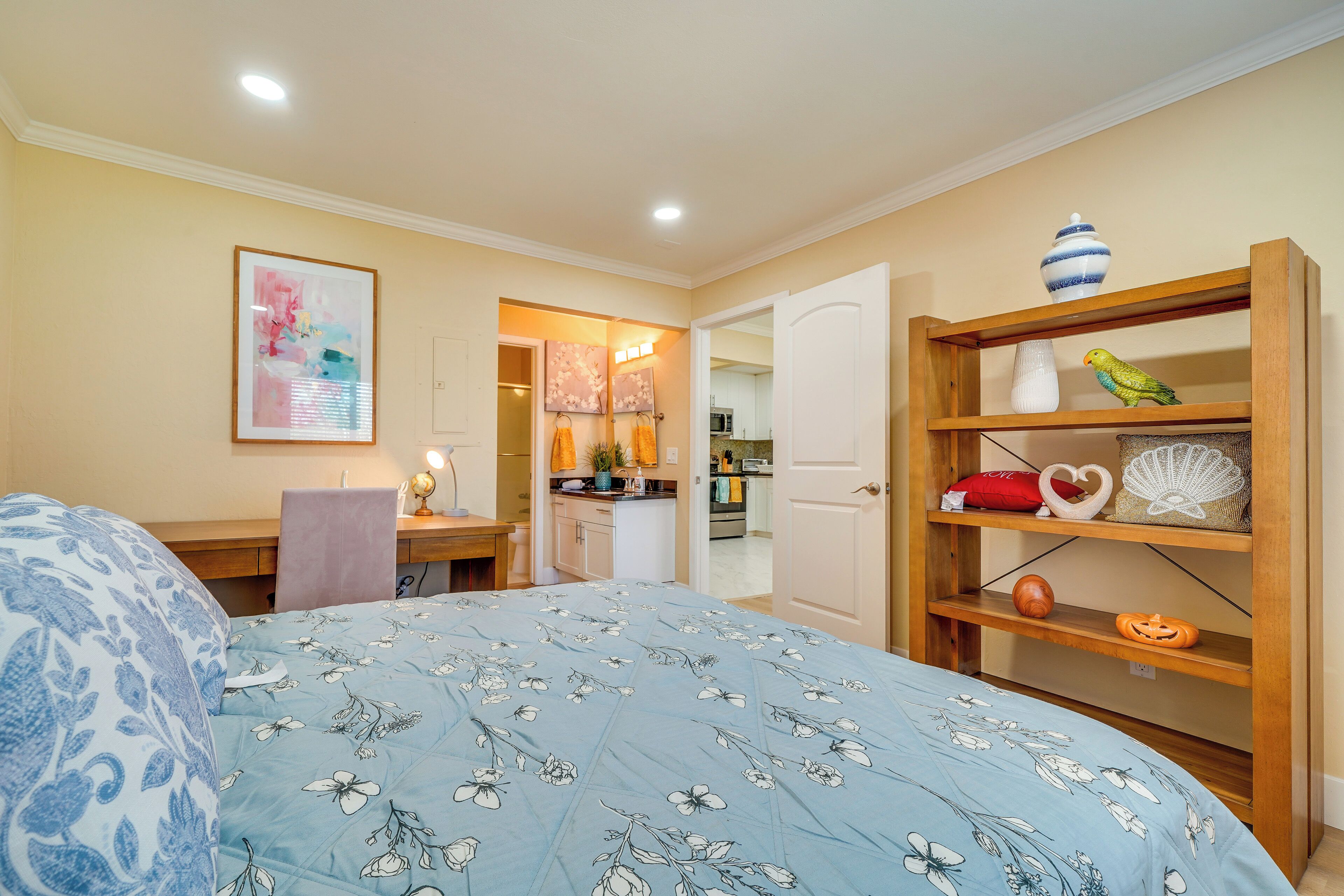 A bedroom with a bed, a desk, a chair, a bookshelf, and a kitchen in the background.