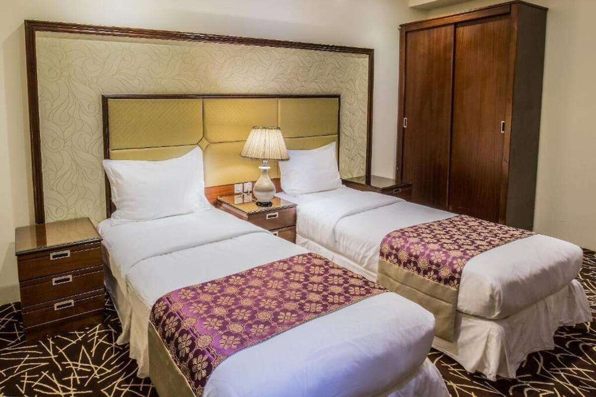 A hotel room with two beds, a nightstand with a lamp, a wardrobe, and a patterned carpet.