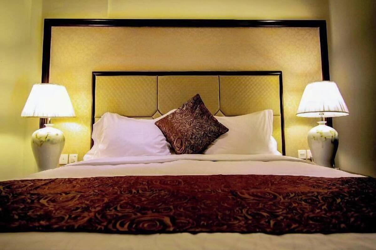 A hotel room with a bed, two bedside lamps, a patterned throw blanket, and a decorative pillow.