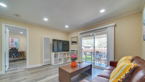 Uma sala de estar com um sofá de couro marrom, uma mesa de centro de madeira com um vaso, uma TV de tela plana e uma porta de vidro deslizante que dá para uma área externa.
