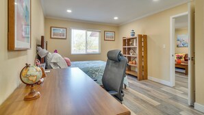 Um quarto com escrivaninha de madeira, um globo, uma cadeira, uma cama com colcha floral, uma janela com persianas e uma estante de livros.