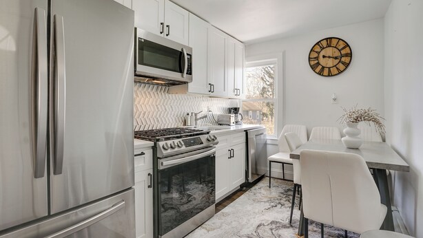 A modern kitchen with stainless steel appliances, white cabinetry, and a dining area with a white table and chairs. - Easy Access to Nyc! Sleek Suburban Apartment (Mamaroneck)