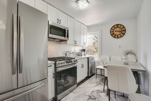 A modern kitchen with stainless steel appliances, white cabinetry, and a dining area with a white table and chairs. - Easy Access to Nyc! Sleek Suburban Apartment (Mamaroneck)