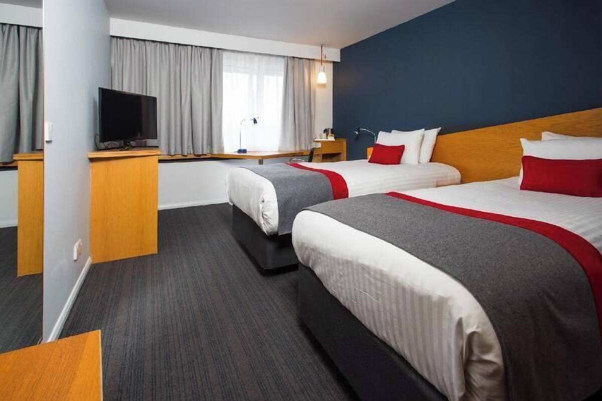 A hotel room with two beds, a desk with a television, a window with curtains, and a dark carpet.
